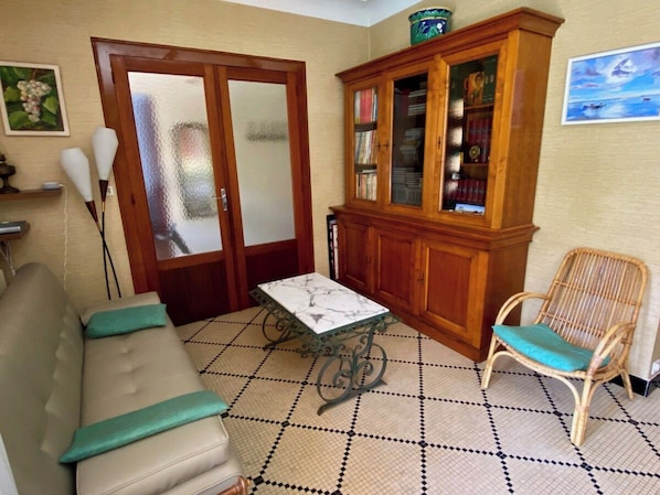 A living room with a sofa, a table, a wicker chair, a cabinet with glass doors, and a patterned floor.