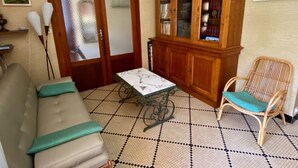Uma sala de estar com sofá, mesa, cadeira de vime, aparador com portas de vidro e piso ladrilhado.