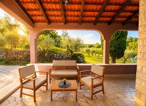 An outdoor patio with a wooden table and chairs, a stone floor, and a view of a green landscape with trees and a stone wall.