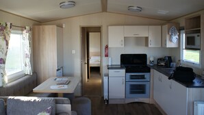 A compact RV kitchen with a stove, microwave, and sink. There is a small dining area with a table and a couch. - Immaculate 3-bed Caravan in Hartlepool (Hartlepool)