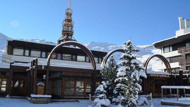 Un bâtiment enneigé, avec une tour de télésiège, une entrée en bois et une enseigne indiquant l’Office de tourisme.