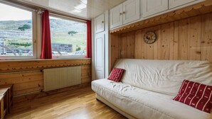 Une pièce avec un plancher en bois, un canapé blanc orné de coussins rouges, une fenêtre avec des rideaux rouges et un mur en bois sur lequel est accrochée une horloge.