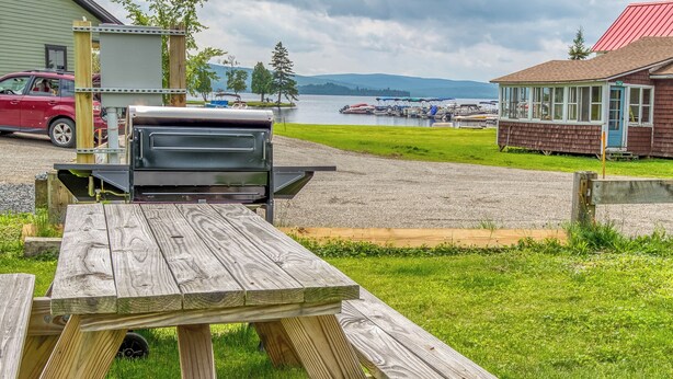 A wooden picnic table and grill in a grassy area with a lake and boats in the background. - 🐾Lakeview on Main - Located in the village of Rangeley with water views! Dog Friendly (Rangeley)