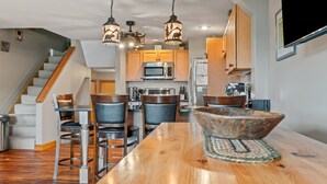 A modern kitchen with wooden cabinets, a stainless steel refrigerator, and a dining area with a wooden table and bar stools. - Town Park Condo D - Great central location in the center of downtown Rangeley (Rangeley)