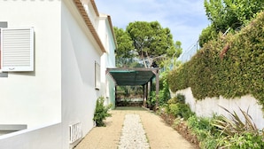 Un étroit sentier de gravier mène à une zone abritée, avec un bâtiment blanc d’un côté et un mur de pierre orné de verdure de l’autre.