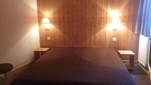 Een kamer met een houten paneel hoofdbord, twee bedlampjes, een bed met een gewatteerde spreideken en een klein houten nachtkastje aan weerszijden.
