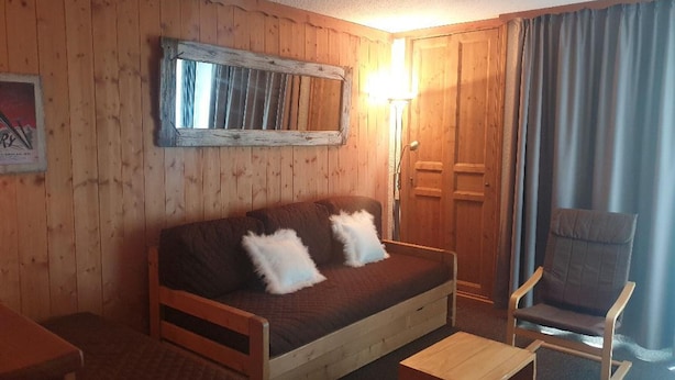 Een gezellige kamer met een houten wand, een bruine bank met twee kussens, een houten fauteuil en een klein houten tafeltje.