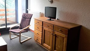Een kamer met een houten dressoir, een televisie erboven, een comfortabele fauteuil en een raam met uitzicht op groen.