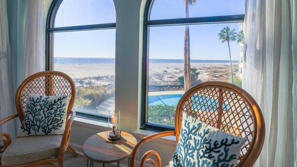 A room with two wicker chairs, each with a cushion featuring a blue coral pattern. A small table with a glass candle holder is between the chairs. Large arched windows provide a view of a beach and palm trees.