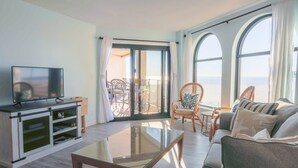 A living room with a TV, sofa, and chairs, a glass coffee table, and large windows with a view.