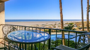 A balcony with a glass table and chairs, overlooking a beach and a pool.