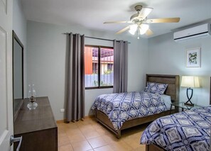 Een slaapkamer met twee bedden, een plafondventilator, een raam met gordijnen, een nachtkastje met een lamp en een airconditioner.