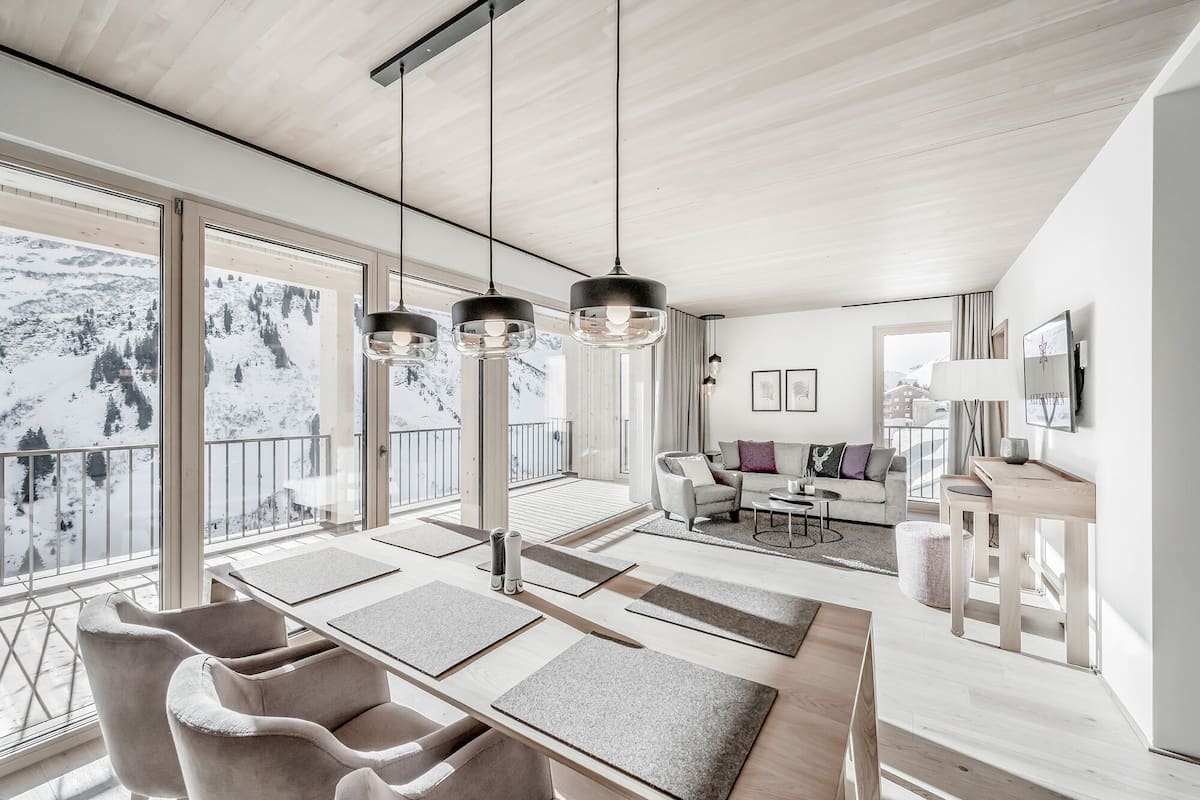 Een ruime woonkamer met een groot raam met uitzicht op een besneeuwd landschap, een eethoek met een houten tafel en stoelen, en een gezellige zithoek met een bank en fauteuils.