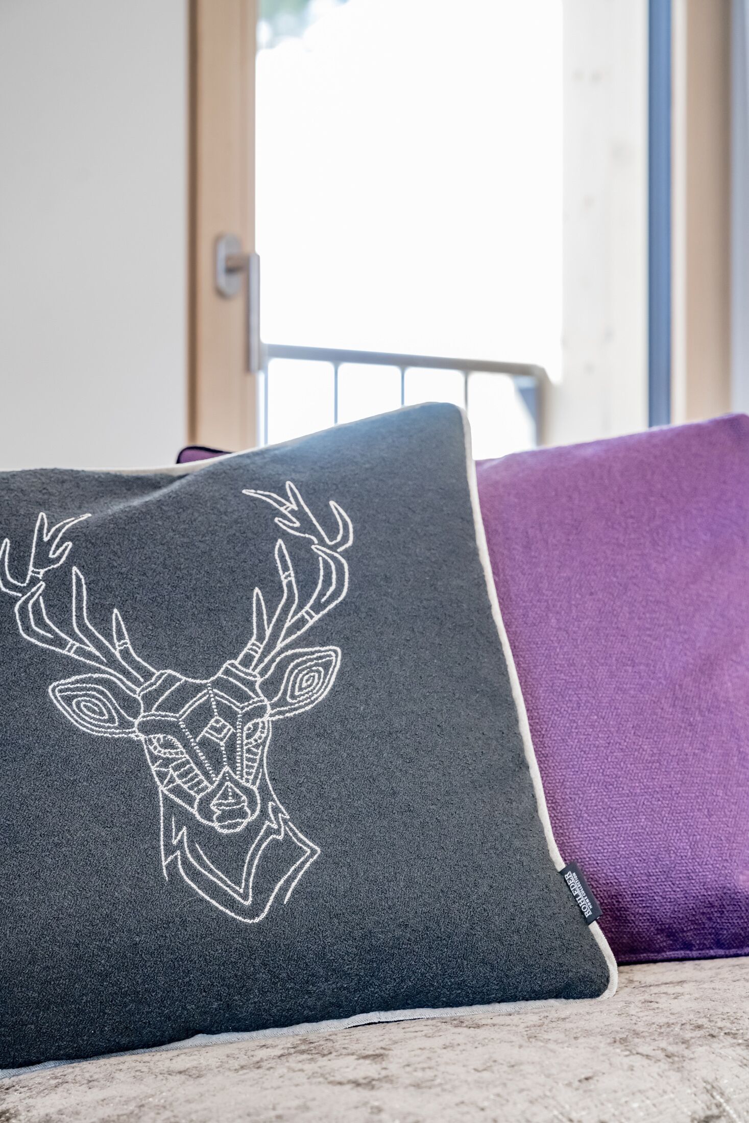 A pillow with a deer design on a dark gray fabric, placed on a couch with a purple cushion.