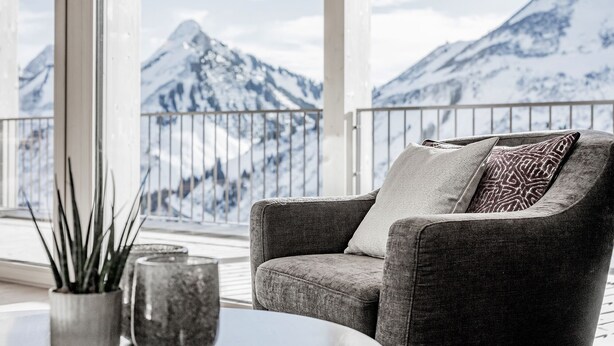 A cozy seating area with a grey armchair and cushions, a round table with two decorative vases, and a view of snow-capped mountains through large windows. - Alpenstern Panoramalodgen (Damüls)