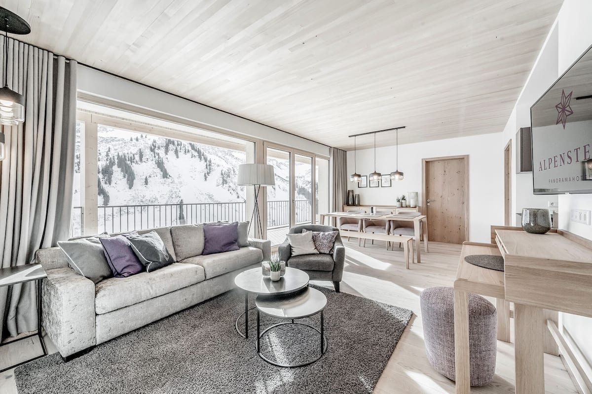 Een moderne woonkamer met een bank, fauteuil en een rond salontafeltje. Er is een eethoek met een tafel en stoelen, en een groot raam met uitzicht op de bergen.