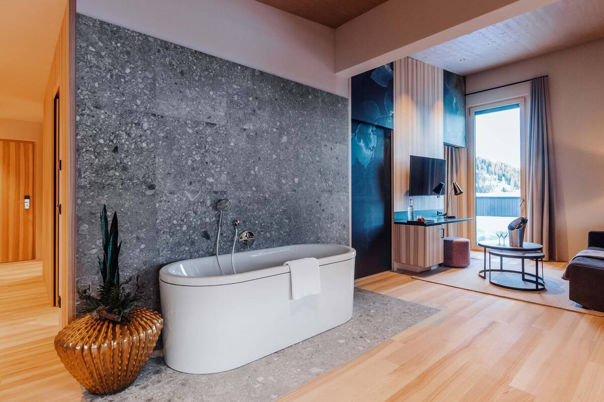 A modern bathroom with a freestanding bathtub, a wooden floor, a large window with a view, and a small seating area.