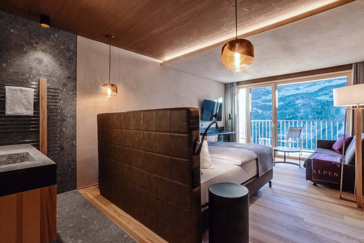 A modern bedroom with a large bed, a small bedside table, a built-in TV, and a balcony with a view of the mountains.