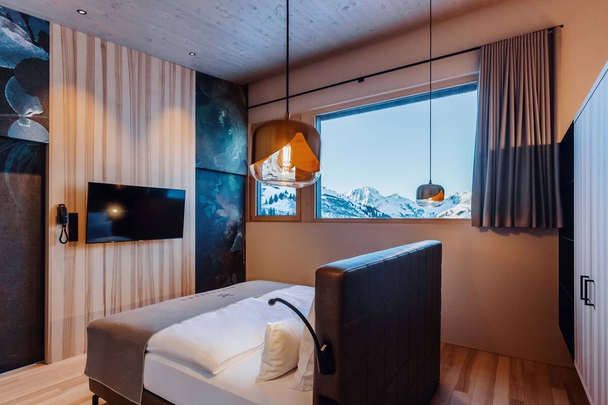 A modern bedroom with a bed, a TV, a window with a mountain view, and a hanging light fixture.