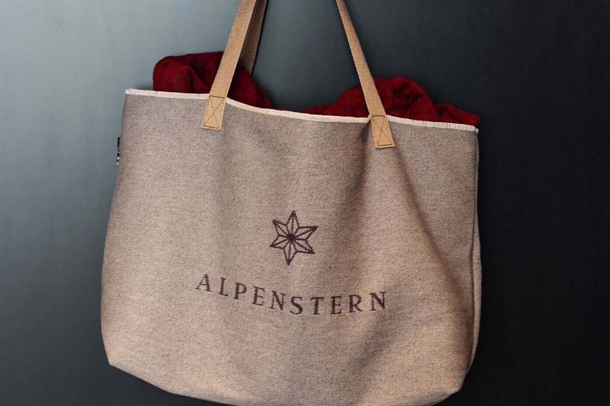 A beige tote bag with "ALPENSTERN" printed on it, hanging on a black wall with hooks.