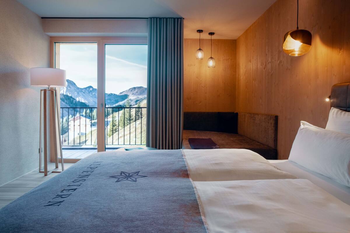 A hotel room with a large bed, a view of mountains through the window, a standing lamp, and a ceiling light.