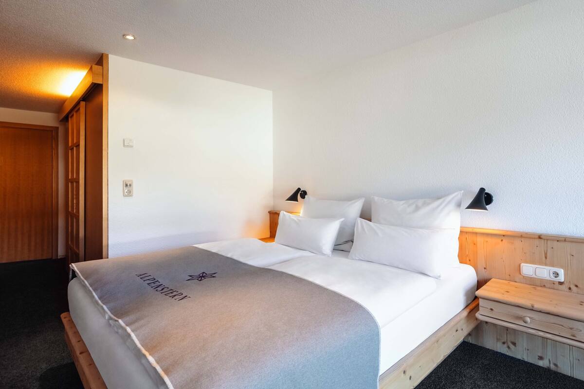 A neatly made bed with a grey blanket, a wooden nightstand with a lamp, and a wooden panel wall.