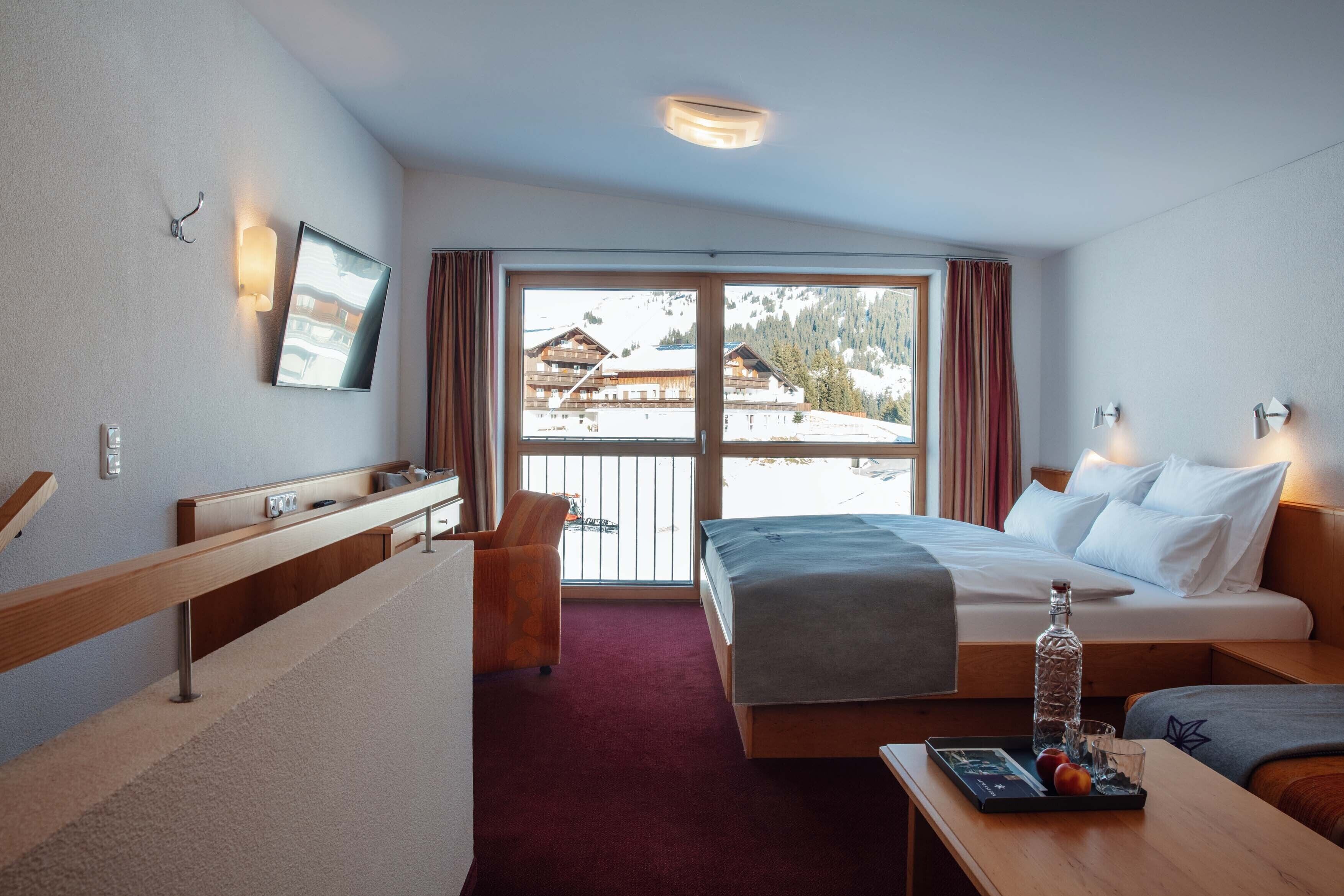 A hotel room with a bed, a desk with a bottle and fruit, a chair, and a large window with a view of buildings and mountains.