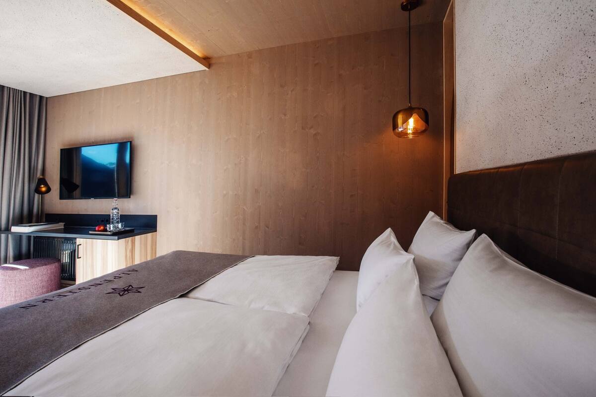 A modern hotel room with a wooden headboard, a flat-screen TV, a bedside table with a lamp, and a large bed with white linens.