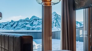 Habitación de hotel con una cama, un ventanal amplio que ofrece vistas a montañas nevadas y una lámpara colgante moderna.