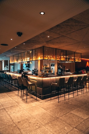 Zona de bar con una barra alargada, varios taburetes y una gran variedad de botellas y copas.