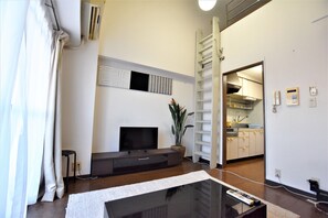 A modern living room with a flat-screen TV, a coffee table, and a potted plant. A kitchen is visible through an open doorway. - Residential Hotel Bevel Tokyo (Tokyo)