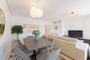 Un salón moderno con una mesa de comedor, sillas tapizadas en gris, un sofá color beige, un televisor de pantalla plana e iluminación colgante.