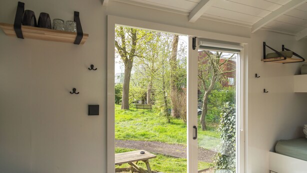 Una habitación con puerta de vidrio corrediza que da a un jardín con mesa de picnic. Hay estantes con objetos en la pared y una cama con colchón verde.