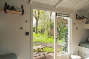 A room with a sliding glass door leading to a garden with a picnic table. There are shelves with items on the wall and a bed with a green mattress. - Tinyhouse Voordek - Lakes of Vinkeveen near Amsterdam (Vinkeveen)
