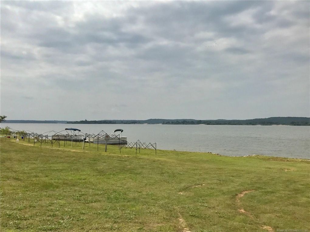 Ein Rasenbereich mit einem Weg zu einem Gewässer, ein umzäunter Bereich mit Stühlen und eine ferne Aussicht auf eine hügelige Landschaft.