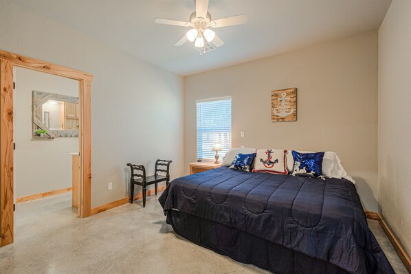 A bedroom with a large bed, a ceiling fan, a window with blinds, a wooden door, and a small table with a lamp.