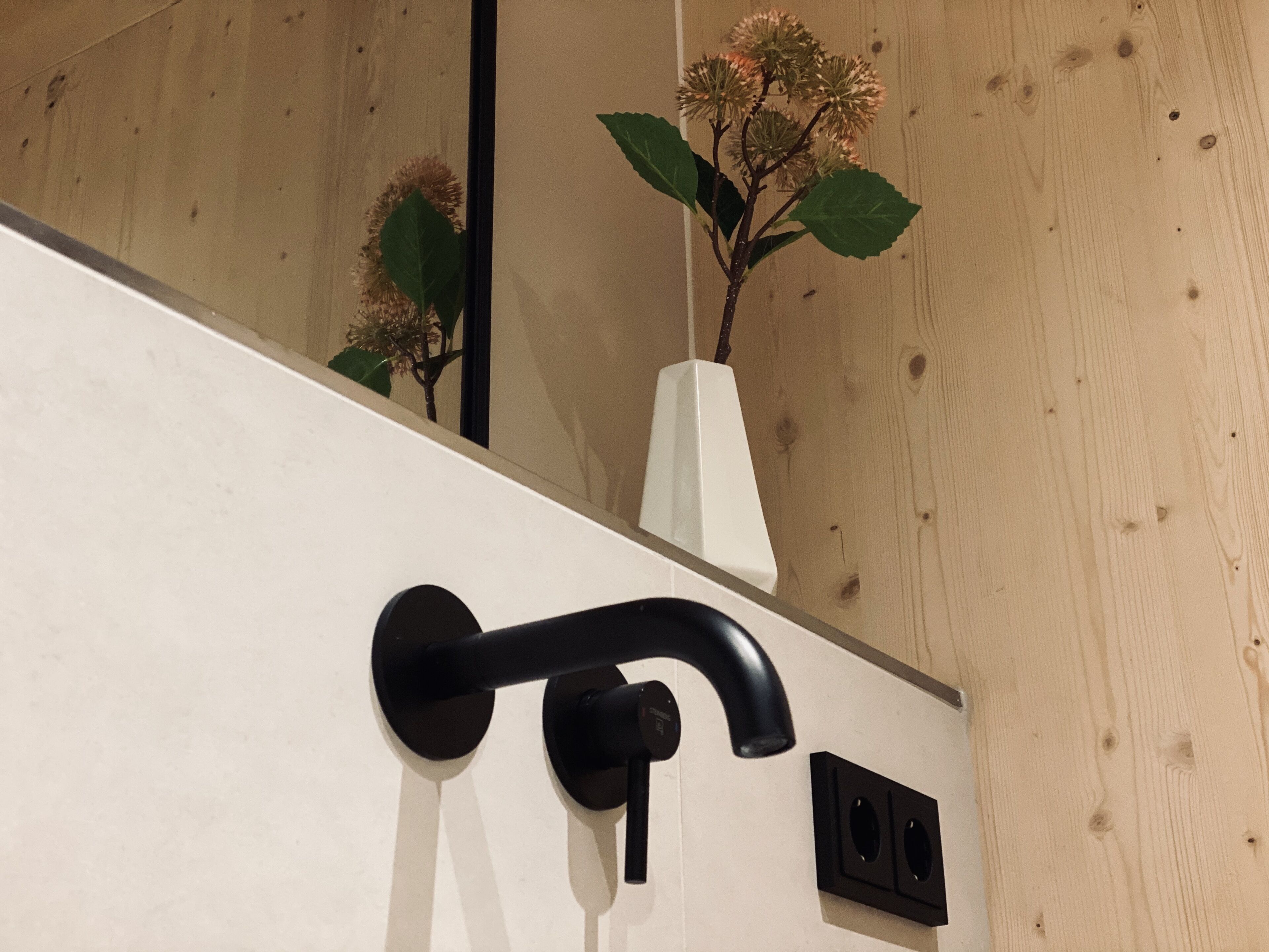 Un bagno con un moderno rubinetto nero, un vaso bianco con fiori e una parete in legno.