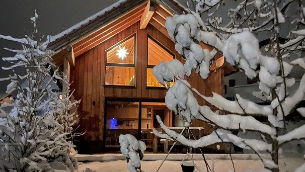 A wooden cabin with a large illuminated window, surrounded by snow-covered trees.