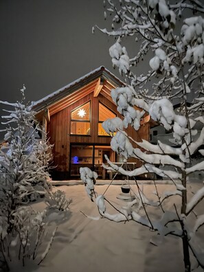 Una cabaña de madera con un ventanal grande iluminado, rodeada de árboles cubiertos de nieve.