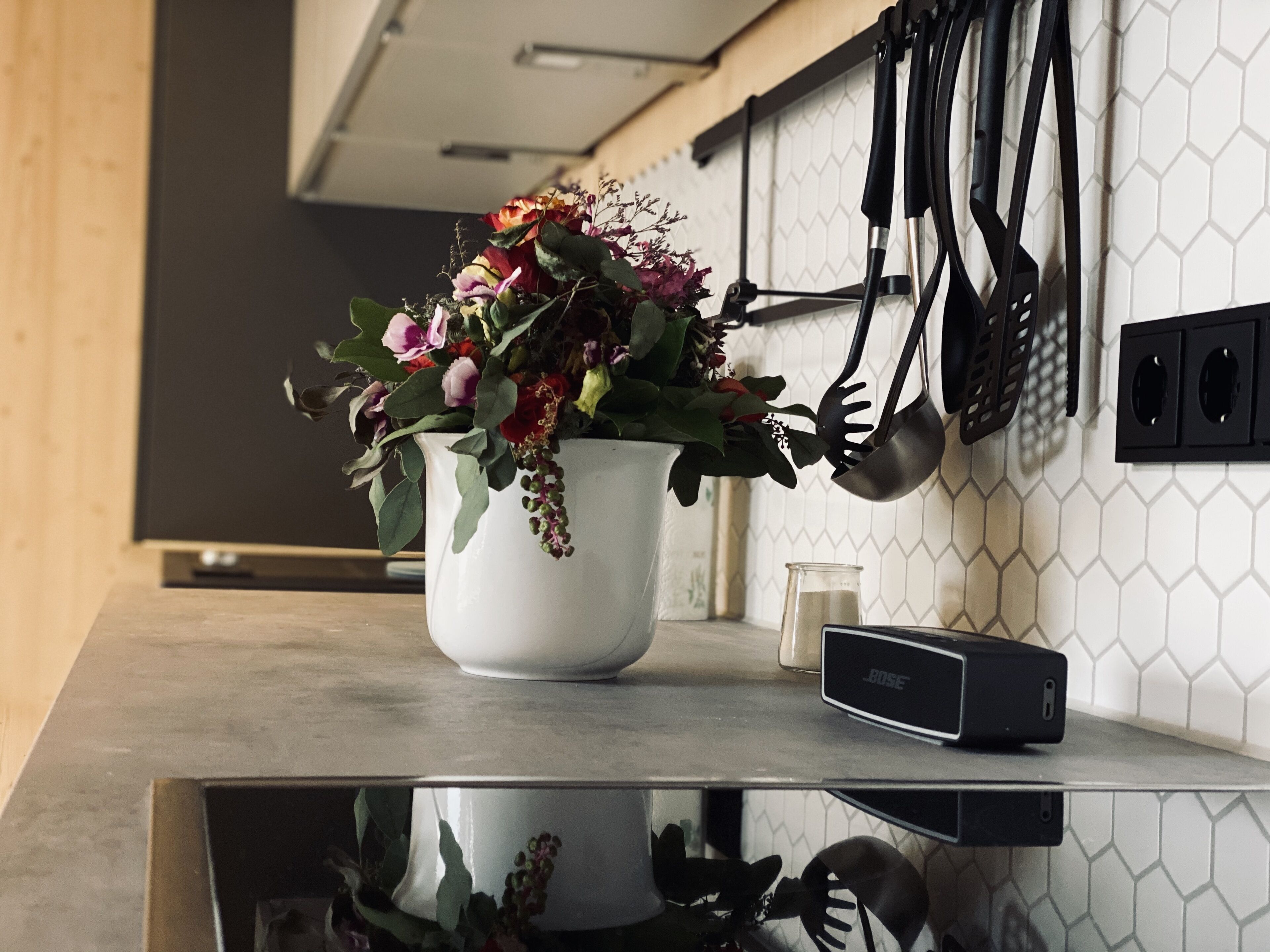 Een aanrecht met een witte vaas met bloemen, een glas water en een zwarte speaker.