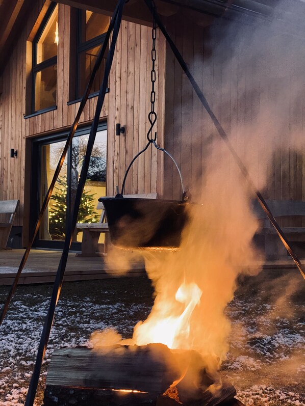 A rustic outdoor fire pit with a hanging pot, surrounded by wooden decking and a wooden building in the background.