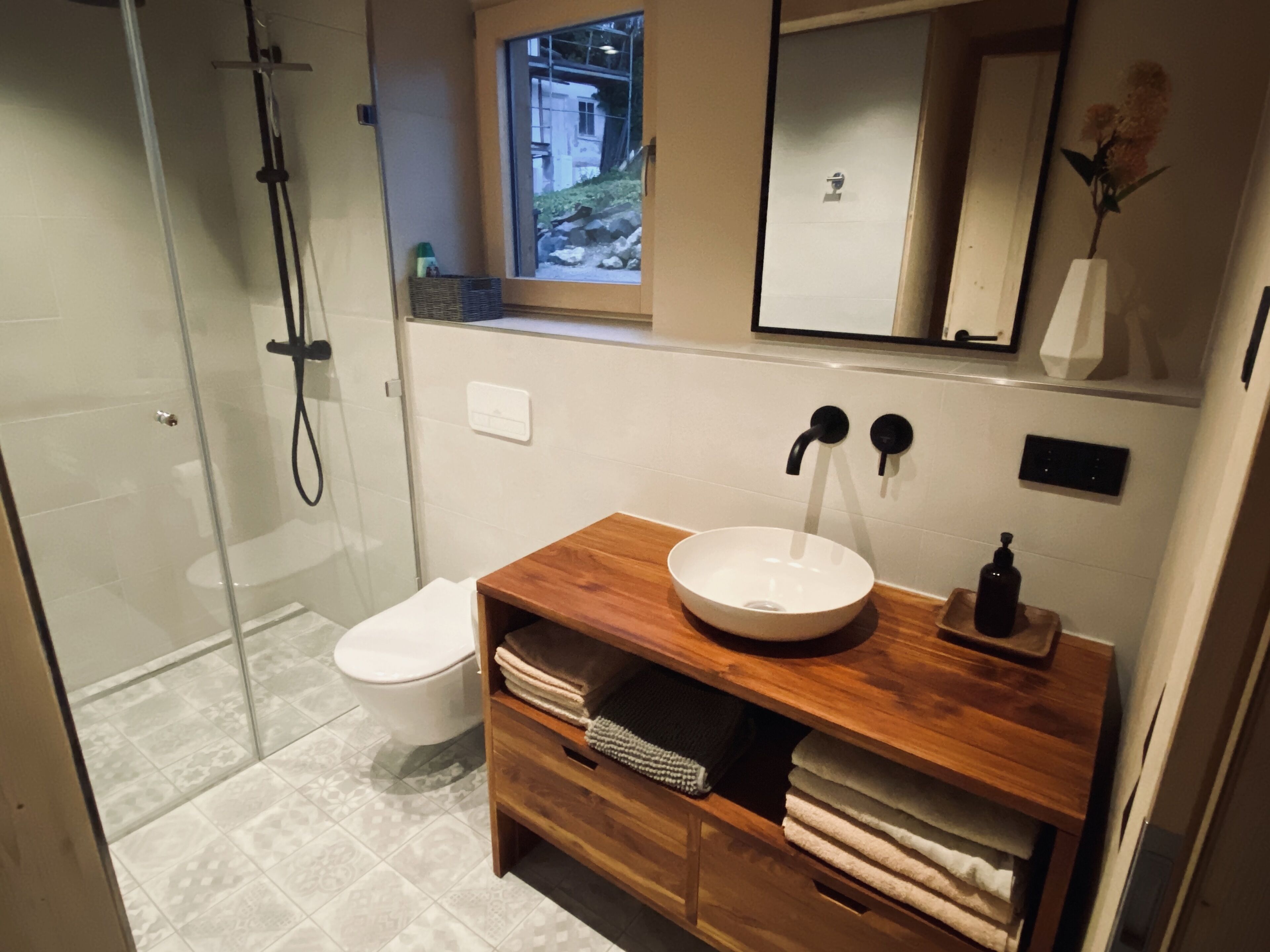 Een moderne badkamer met een douchecabine van glas, een wit toilet, een houten wastafelmeubel met een ronde wastafel en een spiegel erboven.