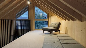 A cozy attic room with a sloped ceiling, large windows, a cushioned bench, and a view of trees.