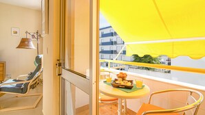 Un balcón con toldo amarillo, una tumbona, una mesita con una bandeja de comida y bebidas, y vistas a edificios.