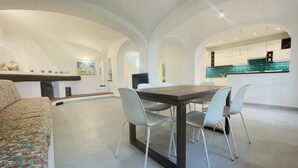 Un espace repas moderne avec une table en bois foncé et quatre chaises blanches, un canapé recouvert d’un tissu à motifs floraux et une cuisine dont le dosseret turquoise est visible à travers une arche.