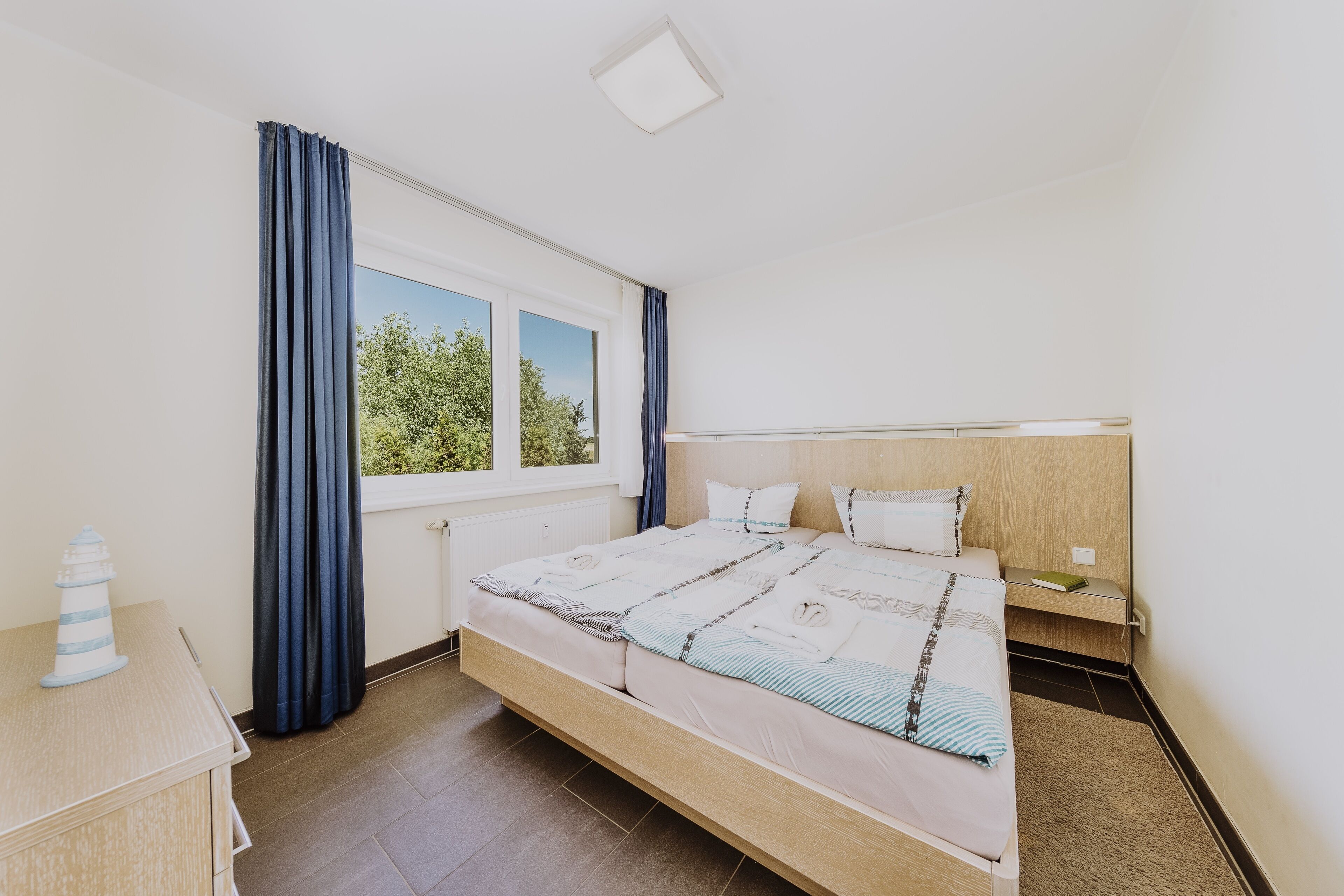 Un dormitorio con una cama grande, una mesita de noche, cortinas en la ventana y vistas a árboles fuera.