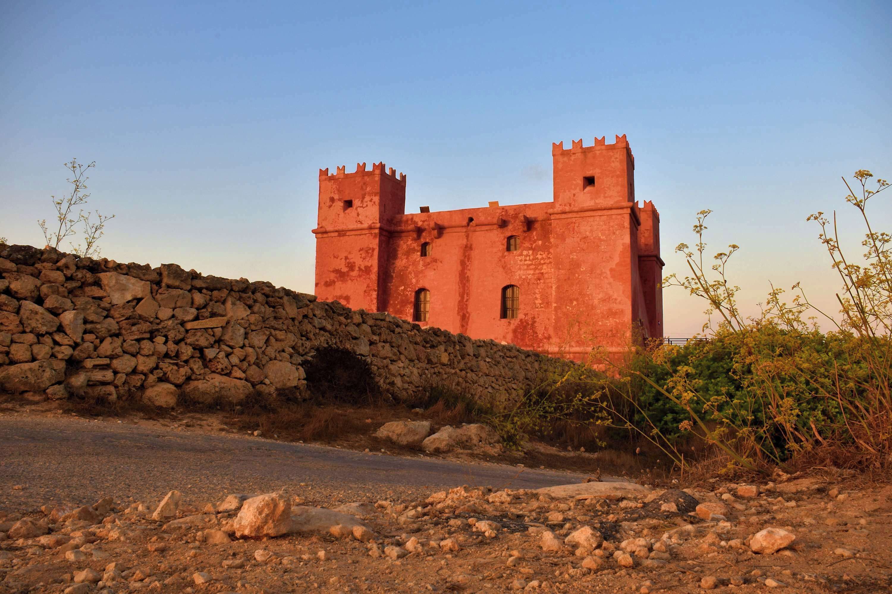 Un gran castillo de piedra roja con dos torres, rodeado de un muro de piedra y vegetación.