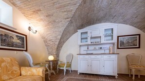 Una habitación acogedora con techo de ladrillo, un mueble blanco con puertas de cristal, un sofá amarillo estampado y dos sillas de mimbre.