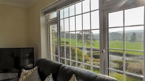 A living room with a black leather sofa, two patterned cushions, and a television. Large windows offer a view of a green landscape. - Bolams Holiday Home (Thomastown)