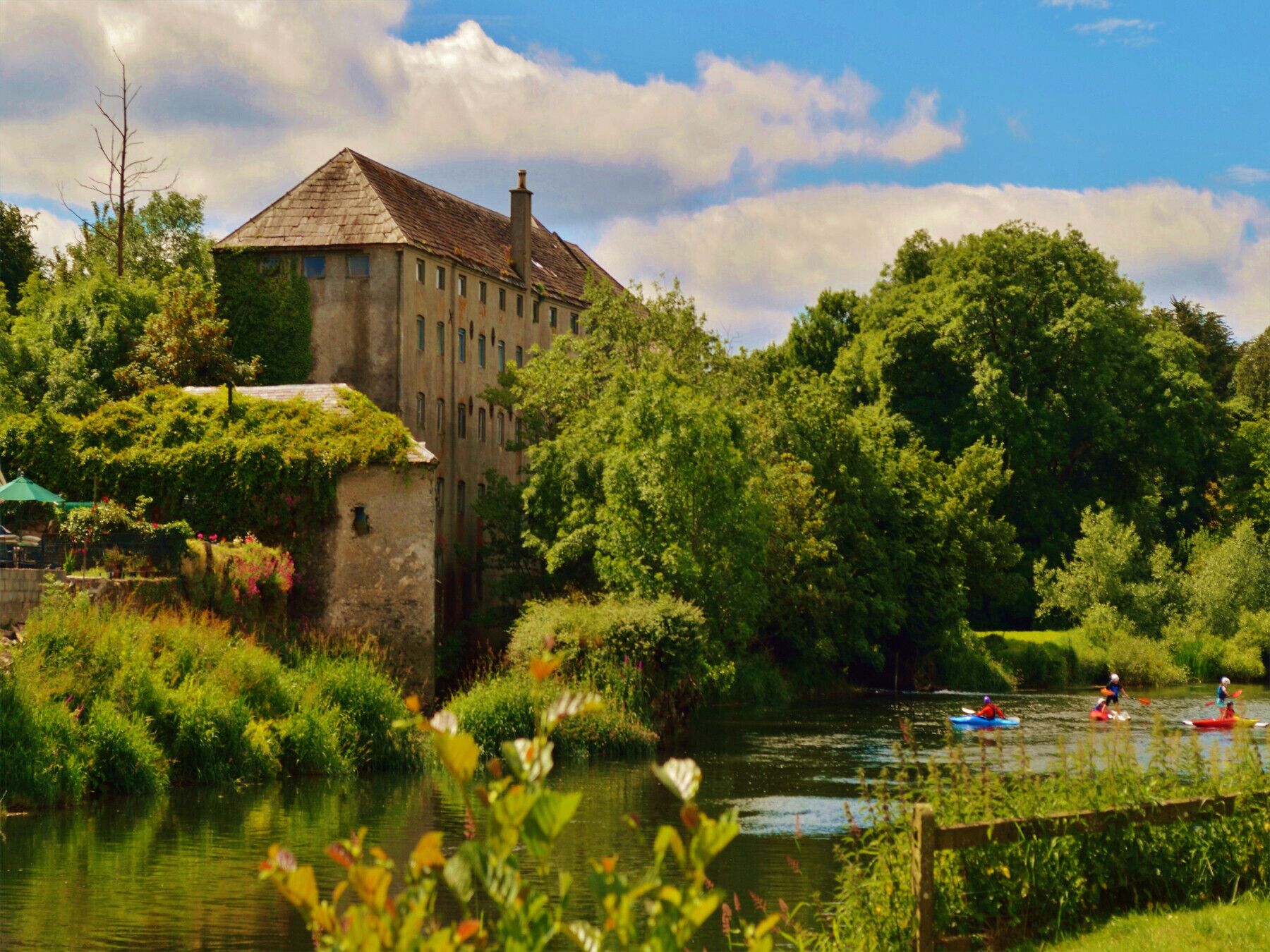 A historic building with a stone facade and a steeply pitched roof, surrounded by lush greenery and a calm river.